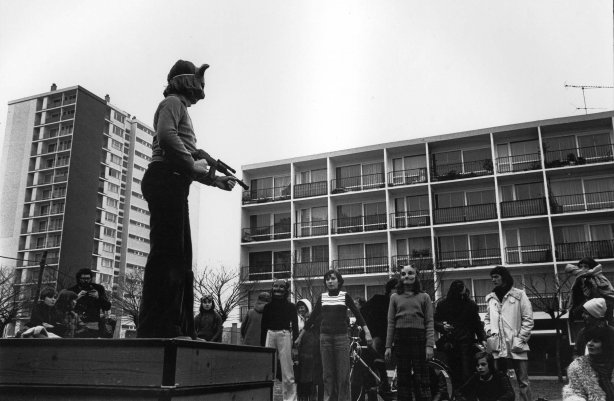 Photographie noir et blanc d'un groupe de jeunes devant 2 barres d'immeubles, l'une des personnes est déguisée avec une arme factice et un masque, les autres la regardent.